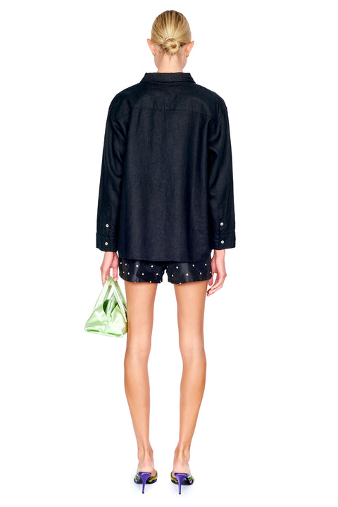 A model shown from the back wearing a black long-sleeve button-up shirt and black shorts with silver studs, holding a metallic green handbag against a plain white background.