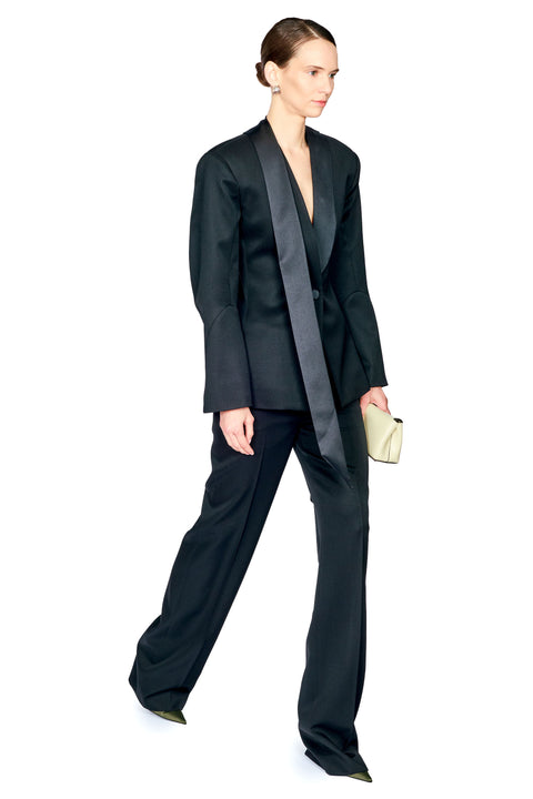 A model wearing a black satin-trimmed blazer and matching wide-leg trousers, holding a small light beige clutch against a plain white background.