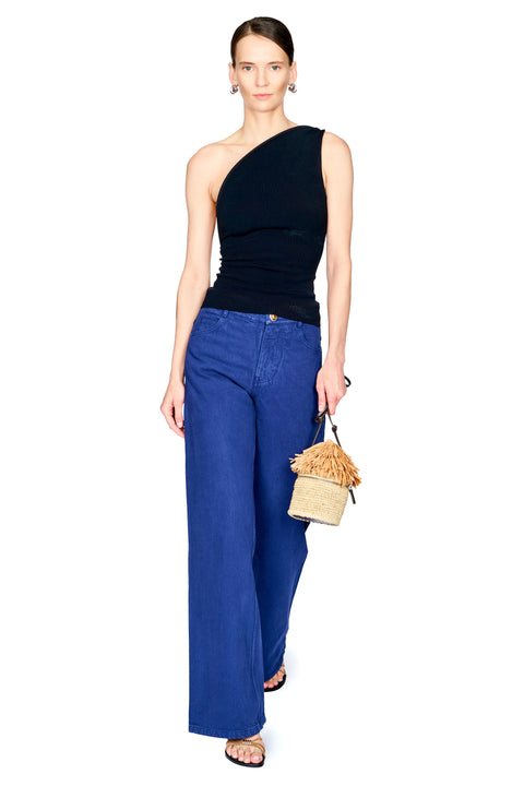 A model stands against a white background wearing a black one-shoulder top, blue wide-leg jeans, and holding a small beige woven handbag.