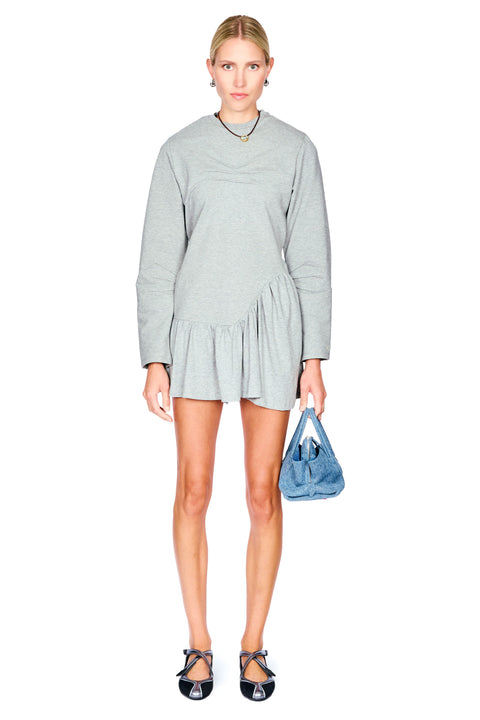 A woman modeling a light gray long-sleeve mini dress with a gathered waist, paired with black strappy heels and holding a blue handbag, against a plain white background.