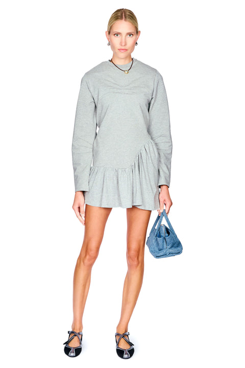 A woman modeling a light gray long-sleeve mini dress with a gathered peplum hem, holding a blue handbag against a plain white background.