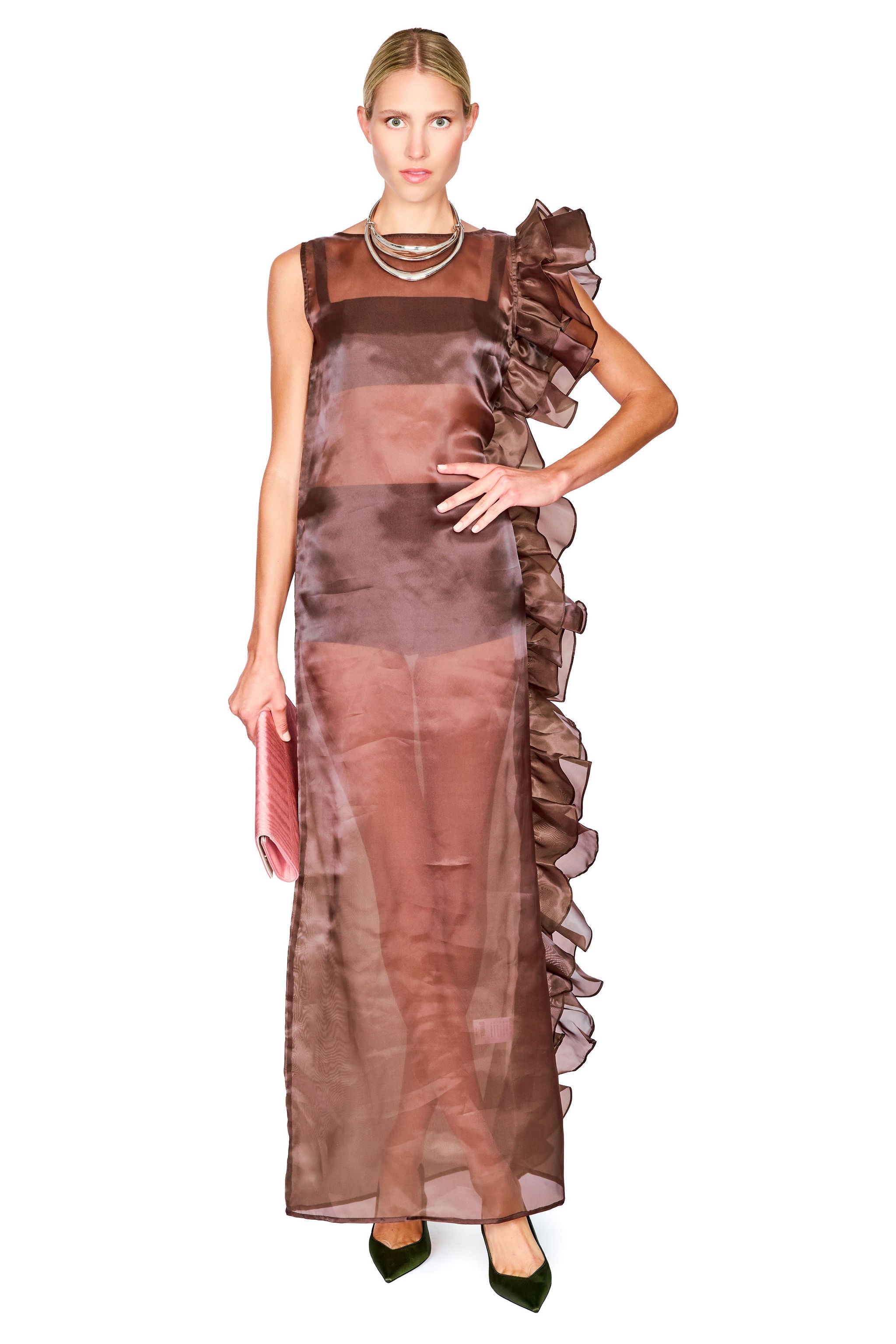 A model wearing a sheer, ruffled brown dress with a high neckline and layered ruffle details along one side, paired with dark green pointed shoes and a pink clutch, set against a plain white background.