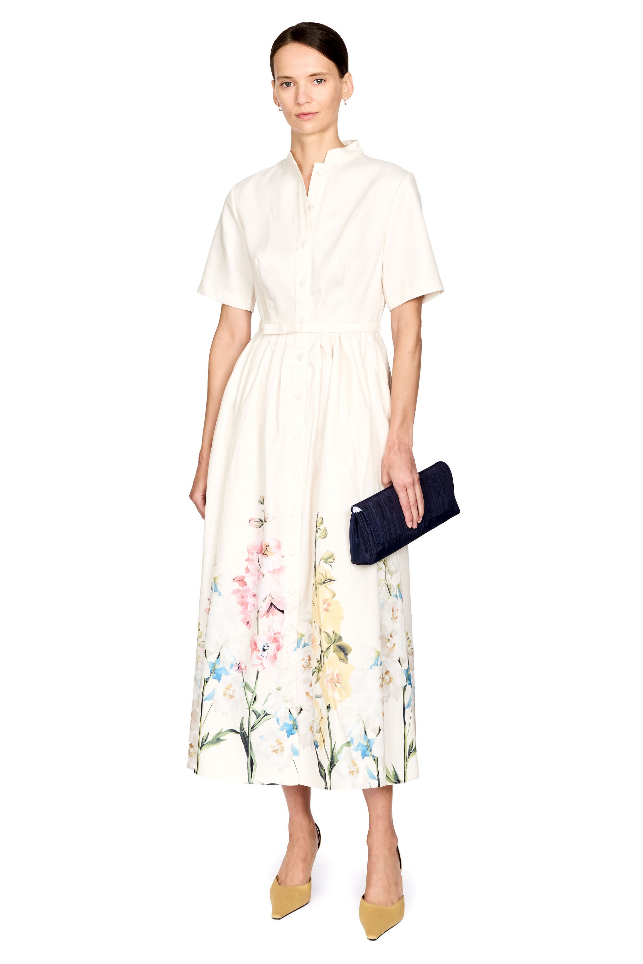 A woman modeling a short-sleeve midi dress with a white base and a colorful floral print concentrated along the hem. She is holding a dark navy blue clutch and wearing yellow heels, set against a plain white studio background.