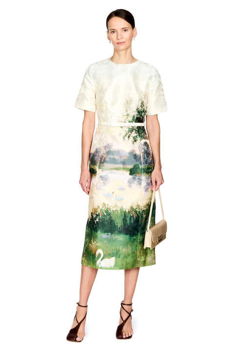A model stands against a white background wearing a short-sleeve midi dress with a cream-colored upper section and a green and blue landscape print on the lower half, featuring a swan near the hem. She is holding a small beige handbag and wearing brown strappy heels.