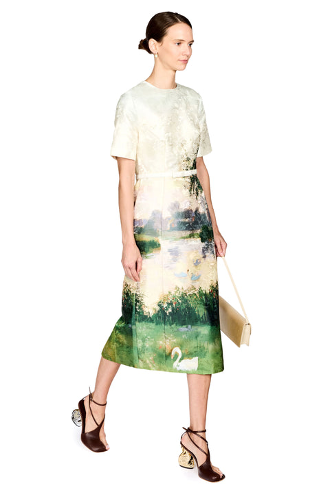 A model wearing a short-sleeve midi dress with a cream lace overlay and a green landscape print featuring a swan, holding a small beige handbag against a white background.