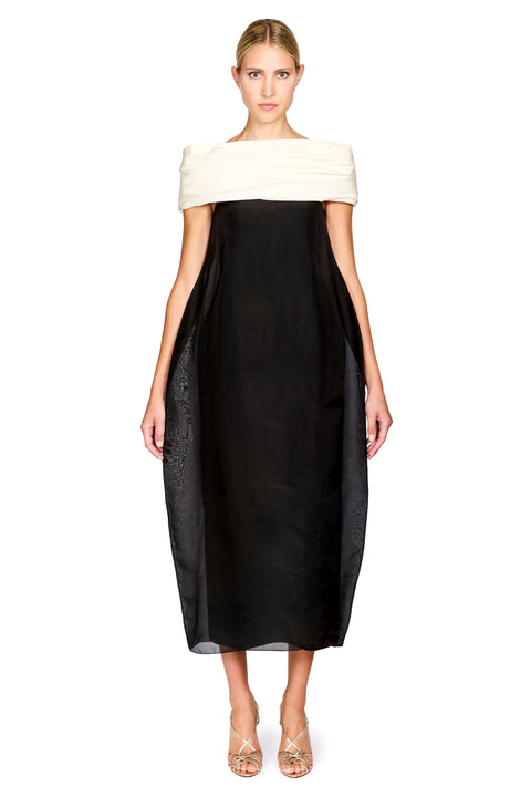A model wearing a black midi dress with an off-the-shoulder design featuring a cream-colored fabric band at the neckline, styled with metallic strappy heels against a plain white background.