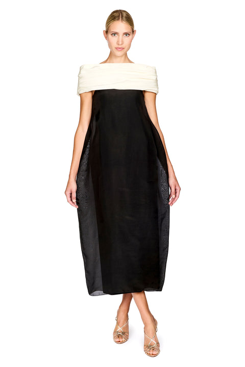A model wearing a black midi dress with a wide off-the-shoulder cream strap and sheer black side panels, paired with clear high-heeled sandals, against a plain white background.