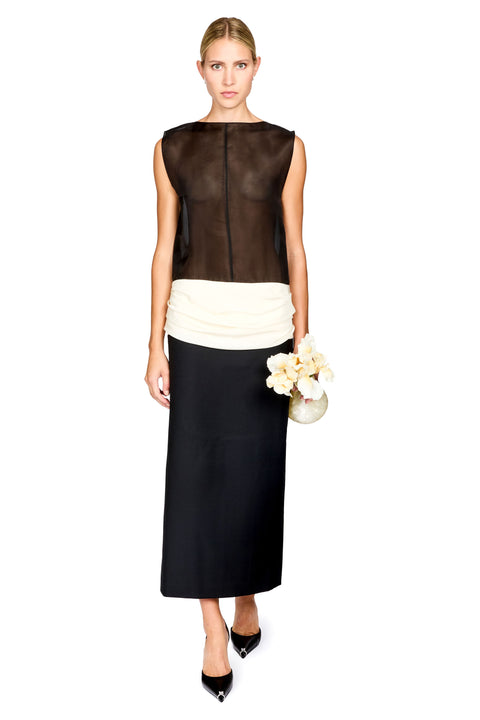 A woman standing in front of a white background wearing a black and white colorblocked long dress with a sheer top and white ruched waistband, holding a flower shaped clutch and black heels