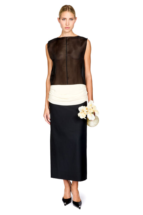 A woman standing in front of a white background wearing a black and white colorblocked long dress with a sheer top and white ruched waistband, holding a flower shaped clutch and black heels
