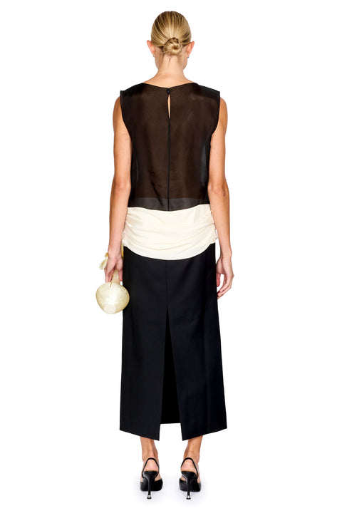 A woman facing the back wearing a black and white colorblocked long dress with a sheer top and white ruched waistband, holding a flower shaped clutch and black heels