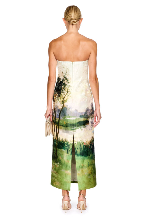 A woman viewed from the back wearing a strapless maxi dress with a scenic landscape print in green, beige, and cream tones. She is standing against a plain white background.