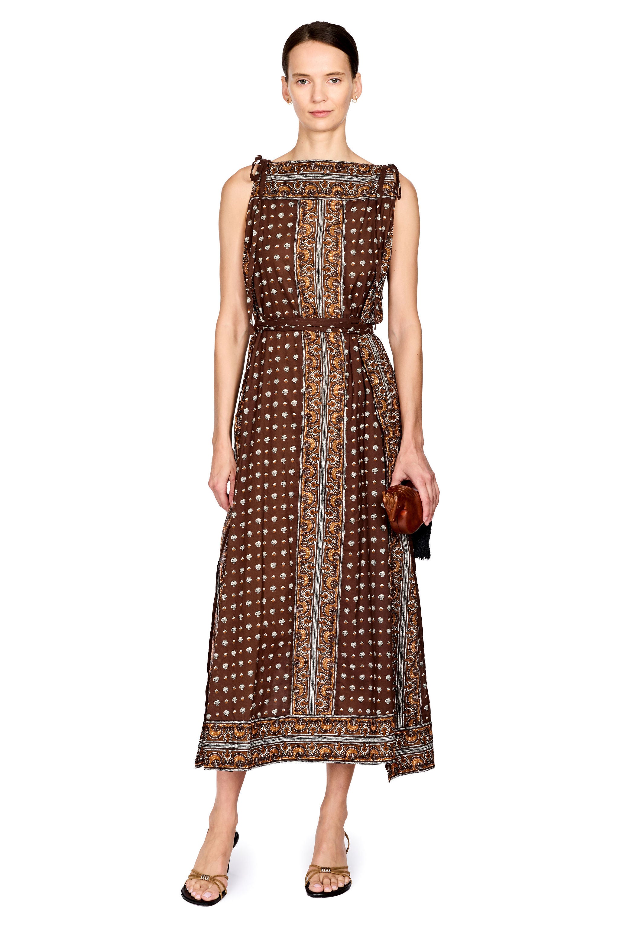 A woman modeling a sleeveless midi dress with a brown base, white polka dots, and vertical orange and blue patterned bands, styled with a matching belt and holding a small brown clutch against a plain white background.