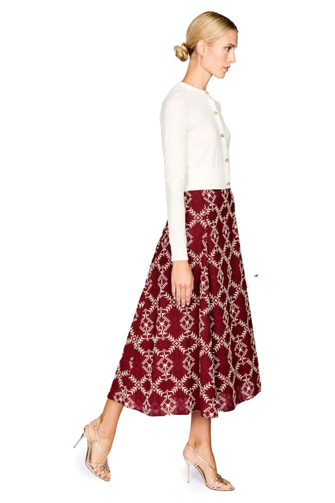 A woman in profile wearing a white long-sleeve cardigan with gold buttons and a burgundy midi skirt with a cream geometric pattern, paired with beige high-heeled sandals, against a solid white background.