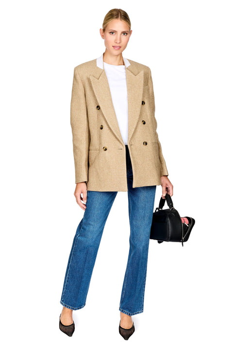 A woman modeling a beige double-breasted blazer over a white top and blue jeans, holding a small black handbag against a plain white background.