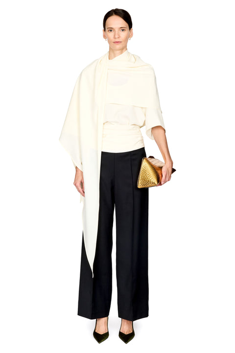 A model stands against a white studio background wearing a cream draped wrap top, black wide-leg trousers, black pointed heels, and holding a textured gold clutch.