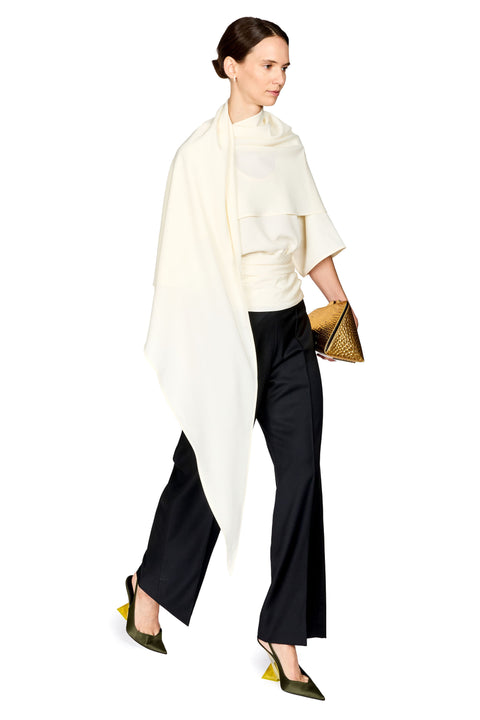 A model is shown from the side wearing a cream draped wrap, a cream short-sleeved top, black wide-leg trousers, olive green pointed-toe heels, and holding a gold textured clutch against a solid white background.