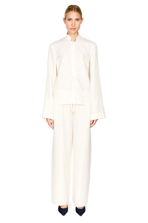 A model stands against a solid white background wearing a matching cream-colored two-piece set consisting of a long-sleeve cropped jacket with a mandarin collar and wide-leg trousers.