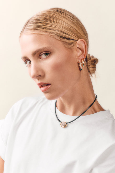 A close-up of a blonde woman with her hair in a low bun, wearing a delicate black cord necklace with a round rose gold pendant and matching rose gold hoop earrings, against a warm off-white background.