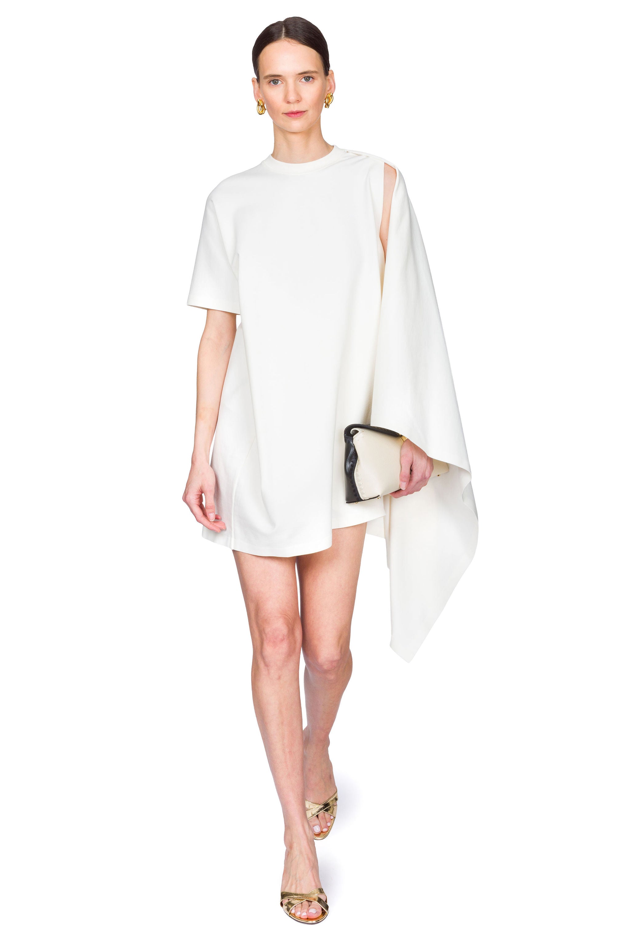 Woman wearing a white dress with a cutout design on one shoulder on a white background clutching a bag in one hand with one foot in front of the other.