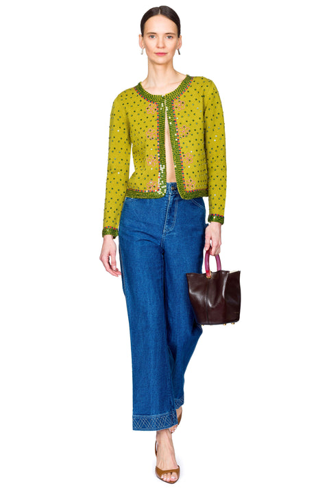 Woman wearing a green patterned top and blue jeans, holding a brown handbag on a white background stepping forward.