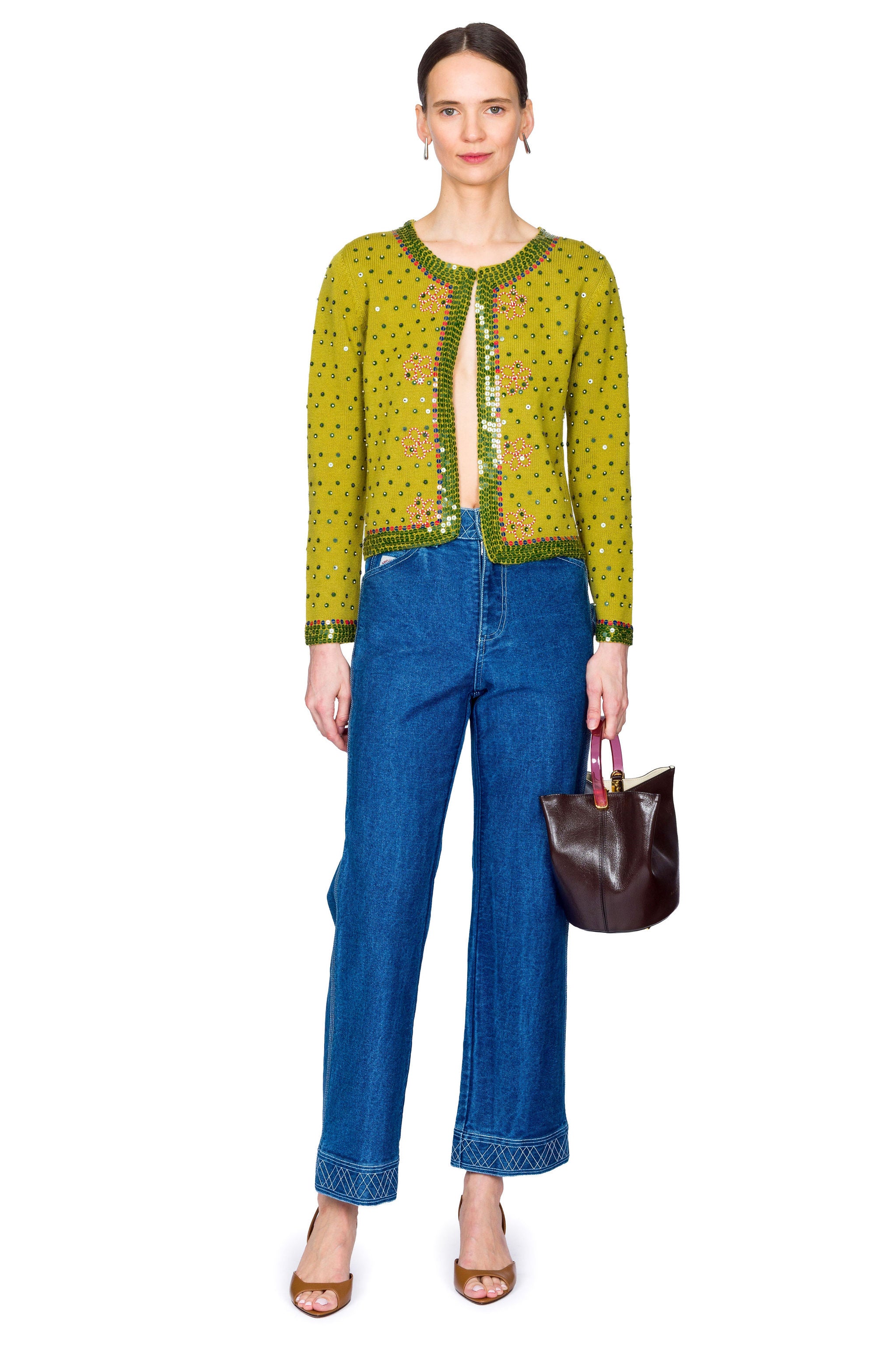 Woman wearing a green embroidered sweater and blue jeans with a brown handbag on a white background with one foot forward.