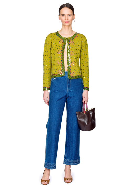 Woman wearing a green patterned top and blue jeans, holding a brown handbag on a white background with one foot in front of the other.