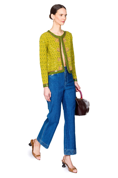 Woman wearing a green patterned top and blue jeans, holding a brown handbag on a white background from the side.