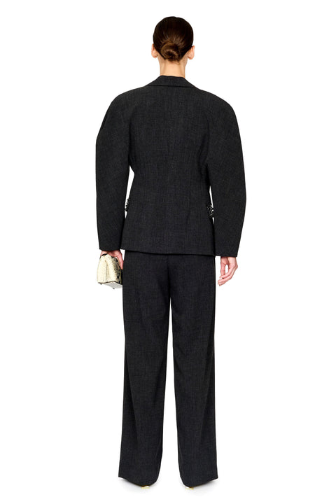 A model shown from the back wearing a dark charcoal gray two-piece suit with a subtle houndstooth pattern, featuring a structured blazer and wide-leg trousers, holding a small metallic clutch against a plain white background.