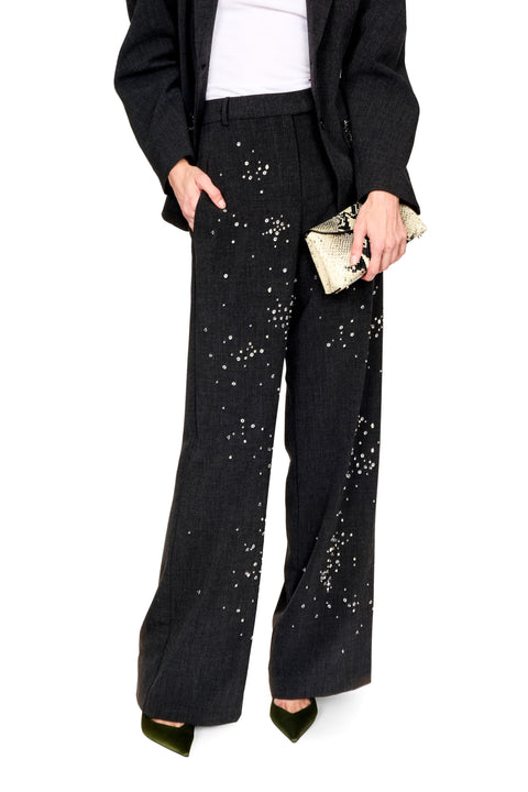 A model wearing black wide-leg trousers embellished with silver rhinestone-like studs, styled with a black blazer, white top, and green pointed shoes, holding a metallic clutch against a white background.