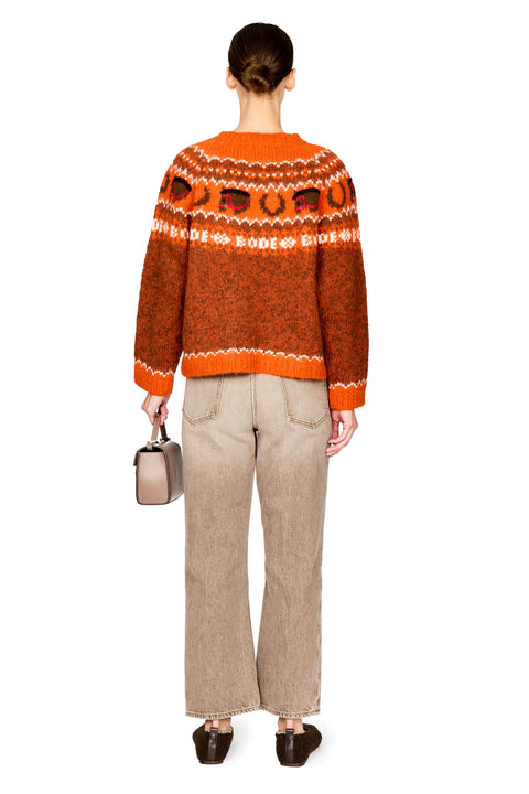 A model shown from the back wearing an orange and brown patterned knit sweater, paired with beige wide-leg trousers and dark brown shoes, holding a small metallic handbag against a white background.