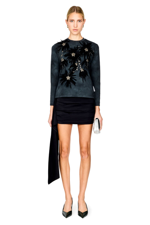 A model stands against a white background wearing a dark charcoal long-sleeve top with black floral embroidery and a black mini skirt paired with black pointed heels and holding a small silver clutch.