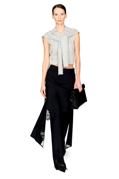 A model wearing a light gray sleeveless cropped top with a large bow detail, black draped trousers with lace inserts, and holding a black clutch bag, set against a plain white background.