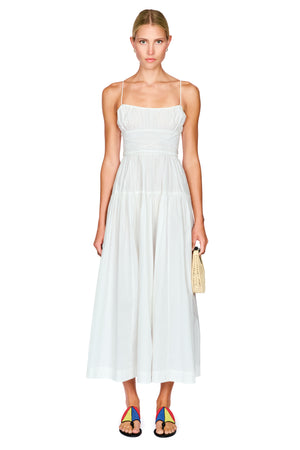A woman modeling a white sleeveless midi dress with thin straps, a gathered bodice, and a flowing skirt, standing against a plain white background and holding a small woven clutch.