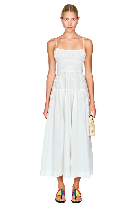 A woman modeling a white sleeveless midi dress with thin straps, a gathered bodice, and a flowing skirt, standing against a plain white background and holding a small woven clutch.