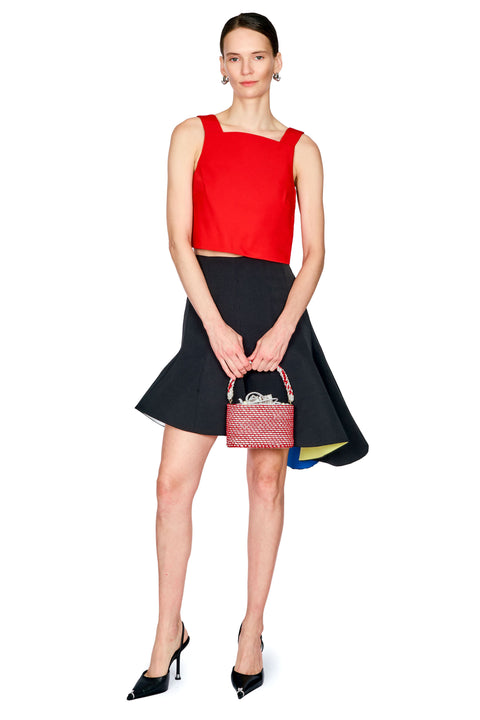 A model stands against a white background wearing a bright red sleeveless crop top and a black flared skirt, holding a small red patterned handbag with black pointed heels.