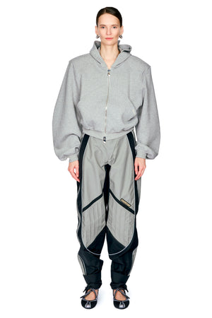 A model wearing a light gray zip-up hoodie and matching gray and black athletic pants, standing against a plain white background.