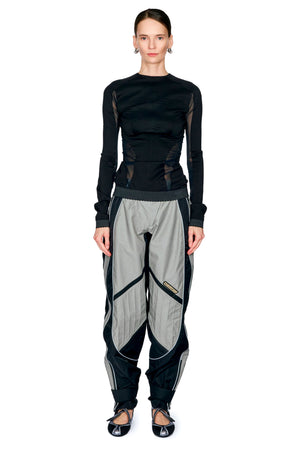 A model stands against a white background wearing a black long-sleeve top with sheer mesh side panels and loose, baggy black-and-gray pants with geometric paneling.