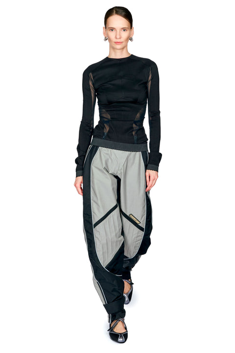 A model wearing a black long-sleeve top with sheer mesh panels and baggy black-and-gray pants, photographed against a plain white studio background.
