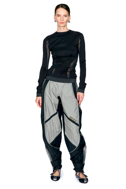 A model wearing a black long-sleeve mesh-paneled top and distinctive light gray and black baggy pants with zippered details, paired with black flats, against a plain white background.
