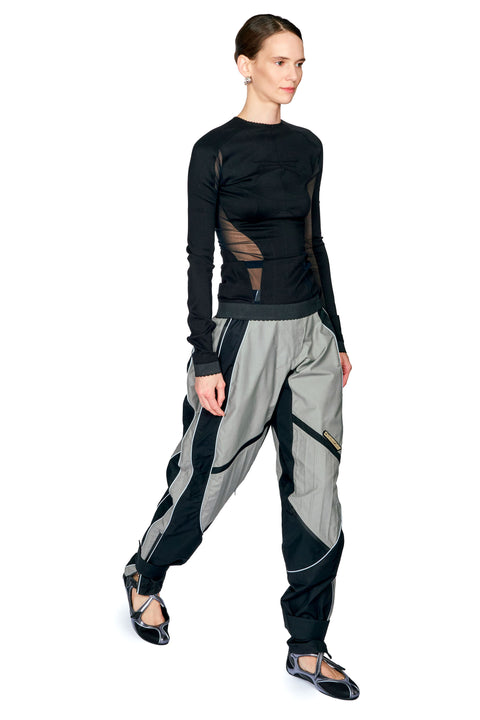 A model wearing a black long-sleeve top with sheer mesh panels and light gray wide-leg pants with black side stripes, posed against a solid white background.