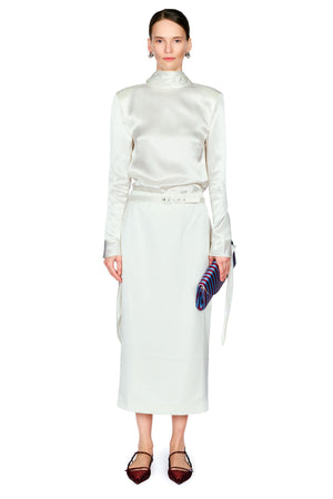 A model stands against a white studio background wearing a matching cream satin long-sleeve mock-neck top and skirt set with a matching belt, holding a purple striped clutch.