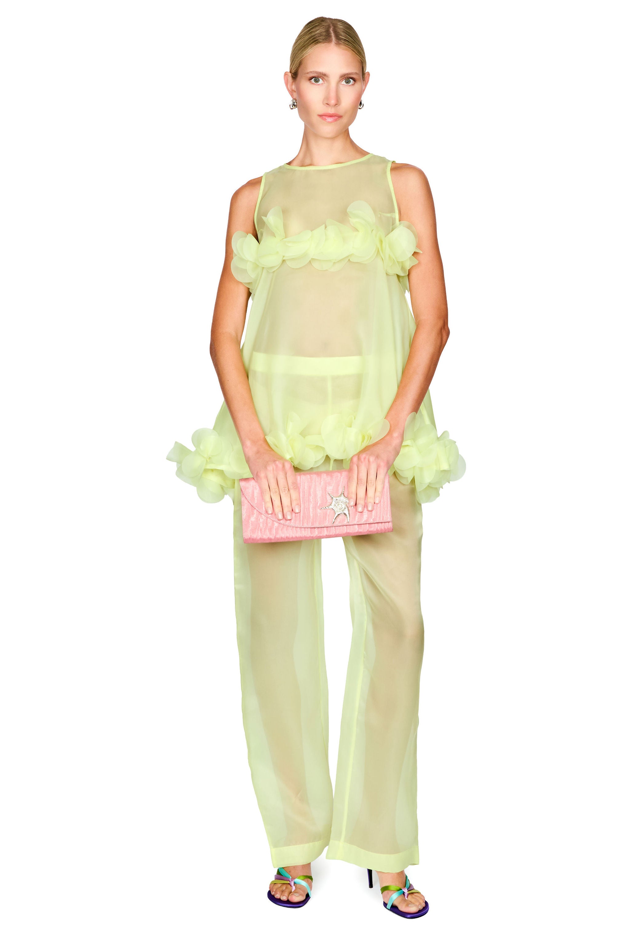 A model wearing a sheer pale yellow-green ruffled sleeveless top and matching wide-leg pants, holding a pink quilted clutch bag, against a plain white background.