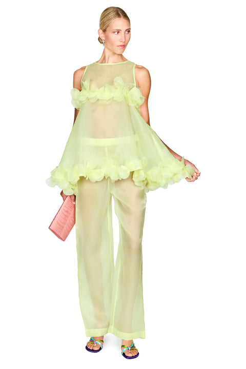 A model wearing a sheer pale yellow-green two-piece outfit with a ruffled halter-style top and matching wide-leg pants, holding a pink clutch against a plain white background.