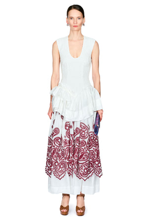 A model stands against a white studio background wearing a sleeveless white top with a ruffled waist and a long white skirt featuring a large burgundy paisley pattern.