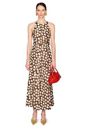 A model stands against a white background wearing a brown halter-neck maxi dress with a cream polka dot pattern, holding a small red handbag.