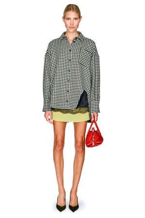 A model stands against a white background wearing a black and white houndstooth button-up shirt, a light green mini skirt, and holding a small red handbag.