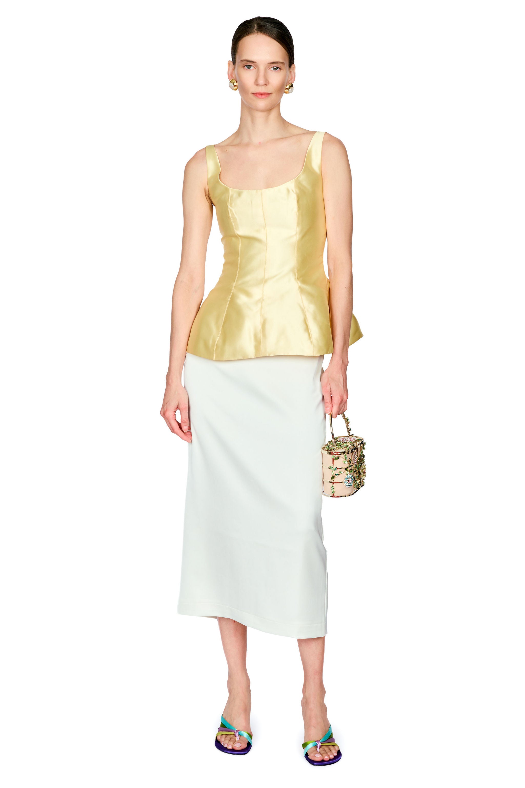 A model stands against a white studio background wearing a sleeveless metallic yellow peplum top and a white midi skirt, holding a small patterned handbag.